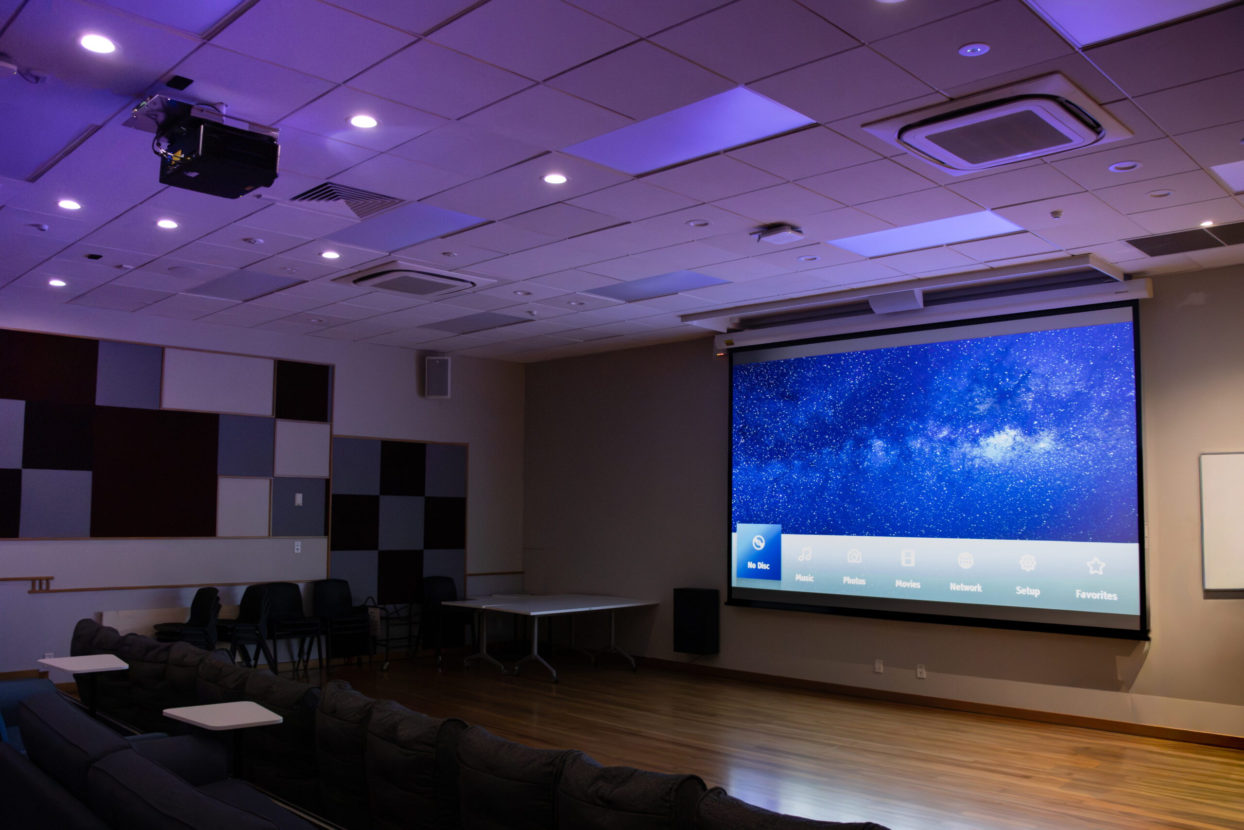 Dimmed cinema room with a large drop-down screen showing a media menu, ambient lighting, and rows of lounge seating.